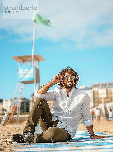 Sommerlicher Lebensstil mit einem jungen dunkelhaarigen kaukasischen Mann mit langem Bart  Sonnenbrille und weißem Hemd an einem Strand. San Sebastian  Baskenland. Im Sand kauernd