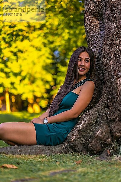 Eine junge hübsche brünette Latina mit langen glatten Haaren lehnt in einem grünen Kleid an einem Baum. Sitzen genießend auf dem Gras neben einem Baum im Park