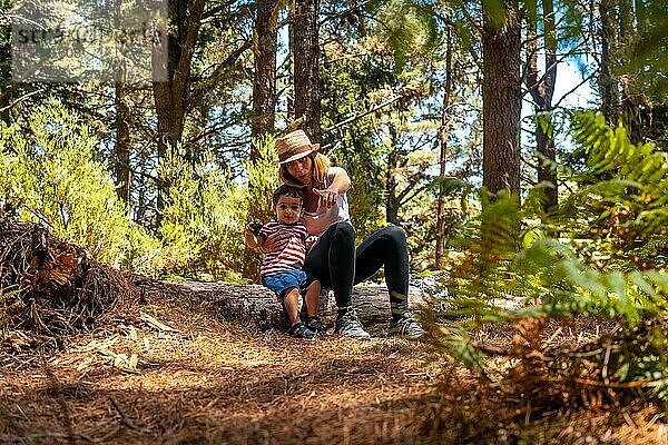 Porträt einer Mutter mit ihrem Sohn  der auf einem Baum in der Natur neben Kiefern sitzt