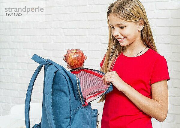 Glückliches Mädchen  das einen Apfel in den Rucksack steckt