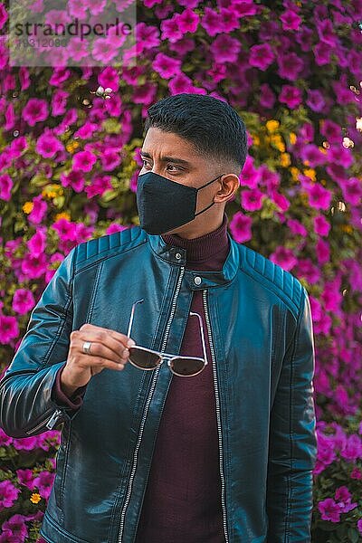 ModeLifestyle Porträt eines jungen Latino mit rosa Blumen im Hintergrund. Jeans  Lederjacke und braune Schuhe. Porträt in der Pandemie mit Maske