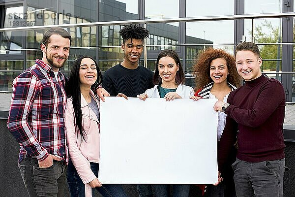 Lächelnde multiethnische Studenten  die ein leeres weißes Plakat halten  das vor einem Glasgebäude steht