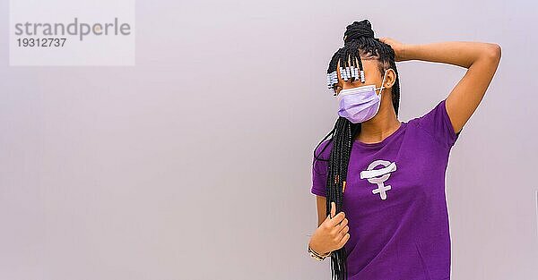 Internationaler Frauentag im Jahr der Coronaviruspandemie Covid 19. Dominikanische Frau mit feministischem lila Hemd und Gesichtsmaske  grauer einfarbiger Hintergrund mit Kopier und Einfügefläche