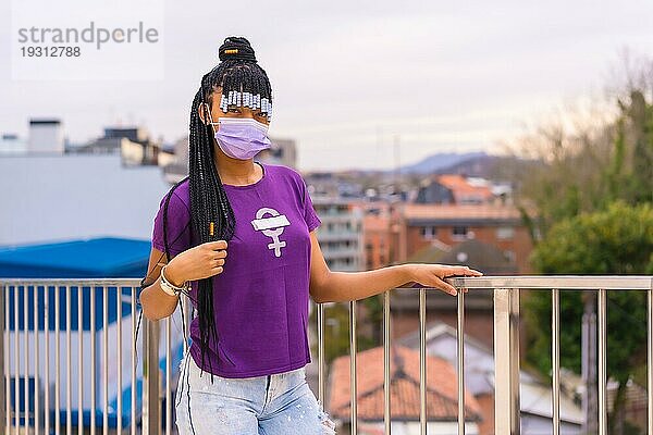 Internationaler Frauentag im Jahr der Coronaviruspandemie Covid 19. Dominikanische Frau mit feministischem lila Hemd und Gesichtsmaske in der Stadt