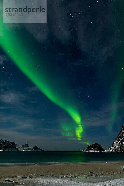 Spektakuläres tanzendes grünes starkes Nordlicht über dem berühmten runden Sandstrand bei Haukland auf den Lofoten in Norwegen in einer klaren Winternacht mit schneebedeckten Bergen