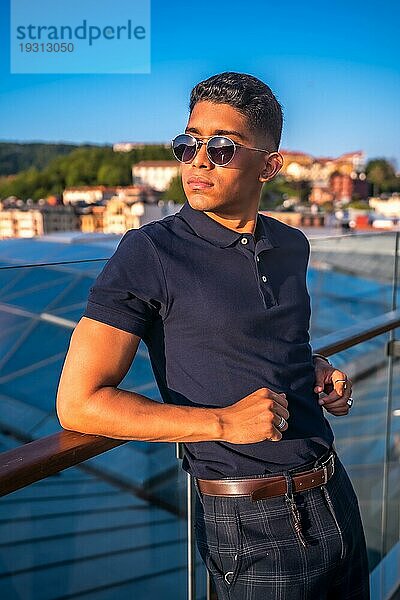 Fashion Lifestyle  Portrait eines jungen Latinos mit der Stadt San Sebastian im Hintergrund  Gipuzkoa. Blaues Polohemd und karierte Hose  den Sonnenuntergang beobachten