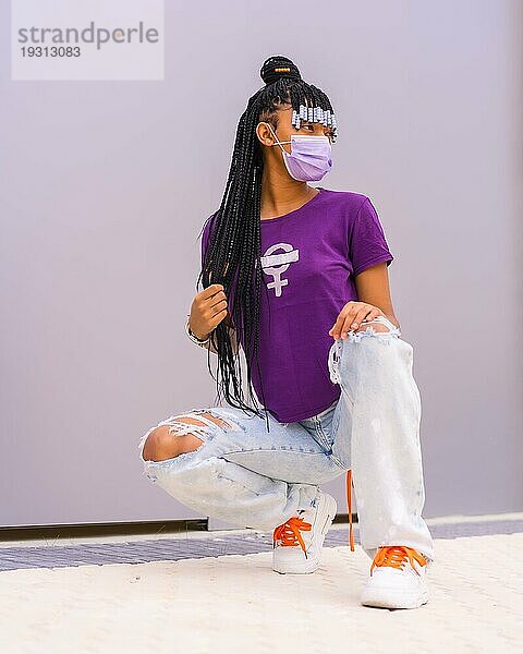 Internationaler Frauentag im Jahr der Coronaviruspandemie Covid 19. Dominikanische Frau mit feministischem lila Hemd und Gesichtsmaske  grauer einfarbiger Hintergrund