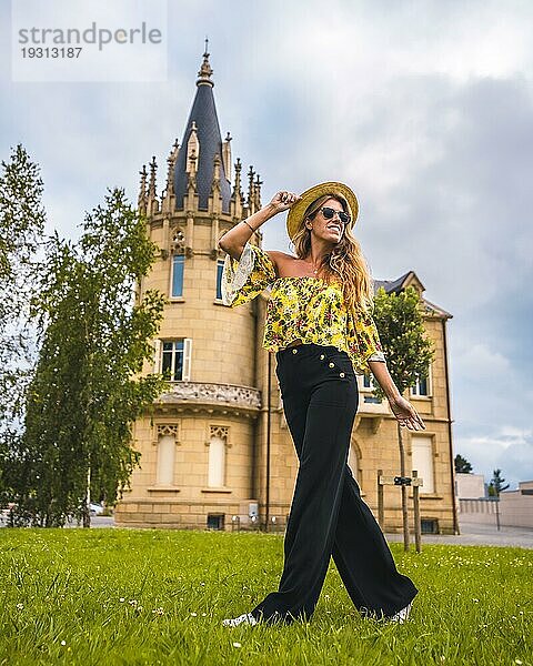 Blonde Frau in einem gelben Vintage Outfit mit Hut geht neben einem Schloss spazieren