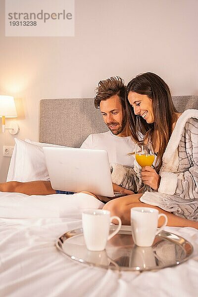 Ein Paar im Pyjama  das beim Frühstück mit Kaffee und Orangensaft im Hotelbett auf den Computer schaut  Lebensstil eines verliebten Paares
