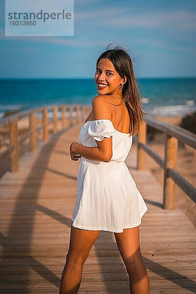 Lebensstil einer jungen brünetten Kaukasierin im Urlaub am Strand  die ein weißes Kleid im Sommer trägt und den Holzsteg entlang geht