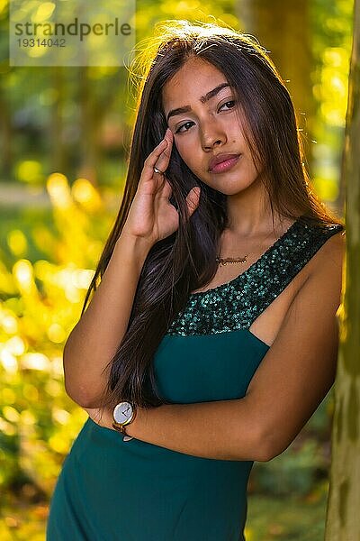 Eine junge brünette Latina mit langen glatten Haaren lehnt in einem grünen Kleid an einem Baum. Porträt mit einem süßen und sinnlichen Blick  vertikales Foto