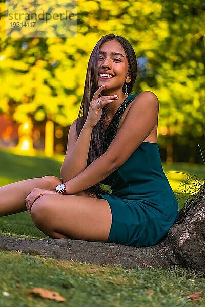 Eine junge hübsche brünette Latina mit langen glatten Haaren lehnt in einem grünen Kleid an einem Baum. Porträt sitzend auf dem Gras neben einem Baum im Park