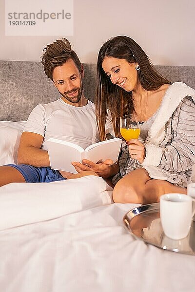 Ein Paar im Schlafanzug liest ein Buch beim Frühstück mit Kaffee und Orangensaft im Hotelbett  Lebensstil eines verliebten Paares. Vertikales Foto