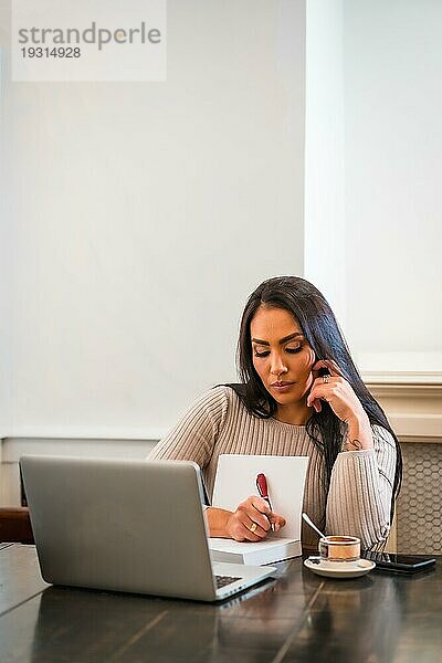 Brünettes Mädchen bei Telearbeit in einer Cafeteria mit Laptop  macht sich Notizen in einer Online Sitzung  vertikales Foto