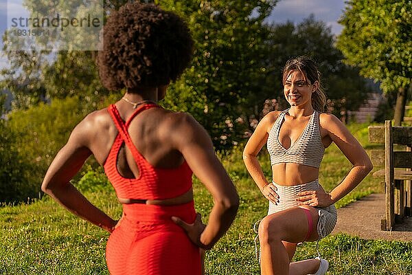 Kaukasisches blondes Mädchen und dunkelhäutiges Mädchen mit Afrohaar machen Stretchingübungen in einem Park. Gesundes Leben  Fitness  Fitness Mädchen  grau und rot Sport Outfits