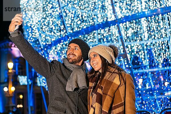 Ein junges europäisches Paar  das die Weihnachtsbeleuchtung der Stadt genießt und ein Selfie mit dem Handy für soziale Netzwerke macht  Lifestyle