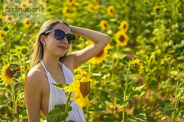 Eine blonde kaukasische Frau mit Sonnenbrille und weißem Kleid in einem schönen Sonnenblumenfeld an einem Sommernachmittag  ländlicher Lebensstil. Navarra  Spanien. Sehr glücklich neben den Blumen