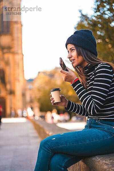 Lifestyle  brünette Kaukasierin  die eine Sprachnachricht in der Stadt sendet. Entspannt mit einem Kaffee zum Mitnehmen neben einer schönen Kirche im Park. Schwarzer Pullover und mit Wollmütze