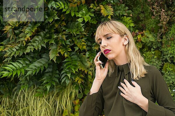 Unternehmerisches blondes Mädchen in einem Gewerbegebiet in einer Ecke der Natur  lächelnd einen Anruf machen  moderner Arbeitsplatz