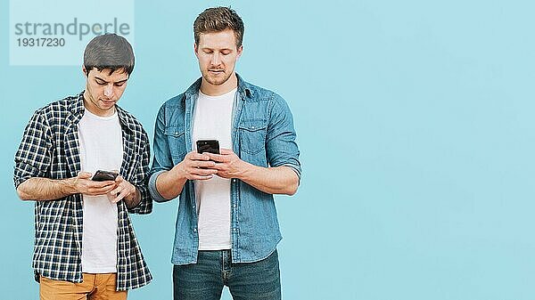 Portrait zwei junge Männer stehen vor blauem Hintergrund und benutzen ein Mobiltelefon