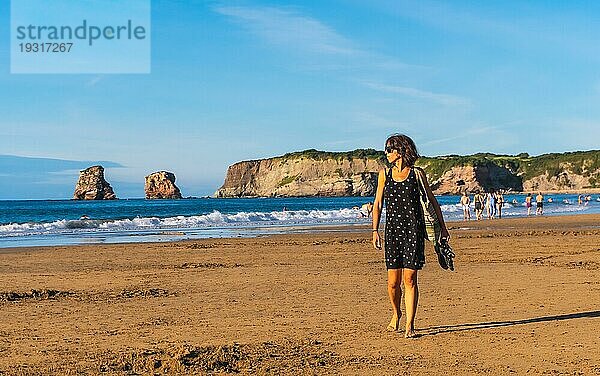 Eine junge Brünette in einem schwarzen Kleid und mit Sonnenbrille geht am Strand von Hendaye im französischen Baskenland spazieren. Frankreich