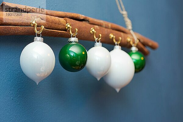 Weiße und grüne Weihnachtskugeln hängen an Haken an einer großen Zimtstange an einer Schnur an einer blau gestrichenen Wand