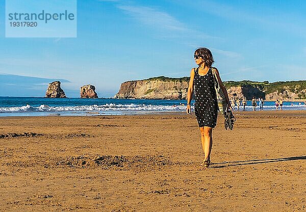 Eine junge Brünette in einem schwarzen Kleid und mit Sonnenbrille geht am Strand von Hendaye im französischen Baskenland spazieren. Frankreich