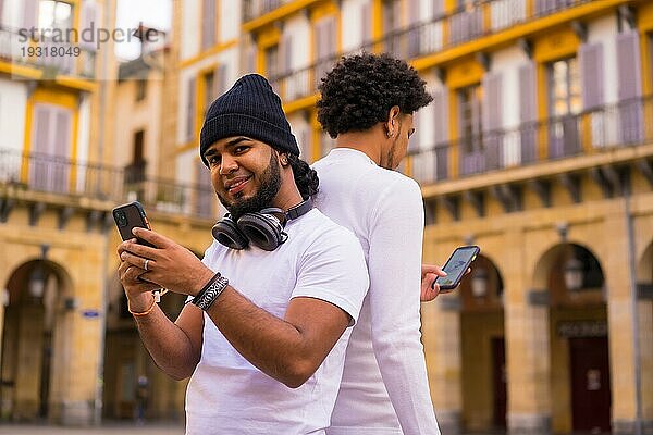 Lifestyle  zwei schwarze Latino Freunde  die Nachrichten versenden oder soziale Netzwerke auf ihrem Handy beobachten. Junge mit Afrohaar und Junge mit Hut und Kopfhörern