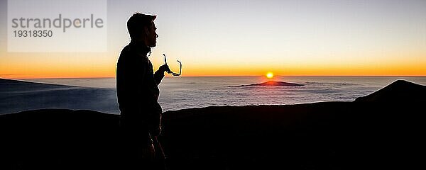 Männliche Person nimmt die Sonnenbrille ab  um den Sonnenuntergang über den Wolken vom Vulkanberg Mauna Kea auf Big Island  Hawaii  zu betrachten