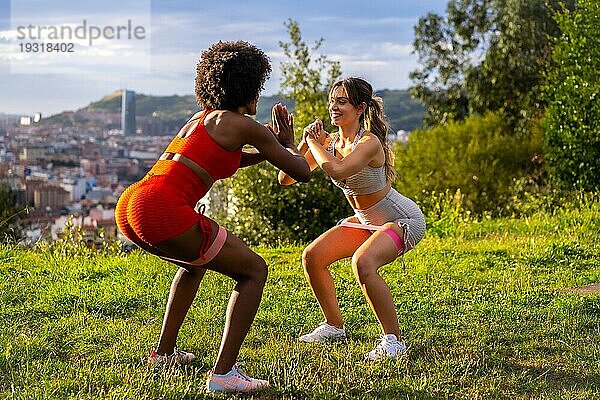 Kaukasisches blondes Mädchen und dunkelhäutiges Mädchen mit Afrohaar tun hocken Übungen in einem Park mit der Stadt im Hintergrund. Gesundes Leben  Fitness  Fitness Mädchen  grau und rot Sport Outfits