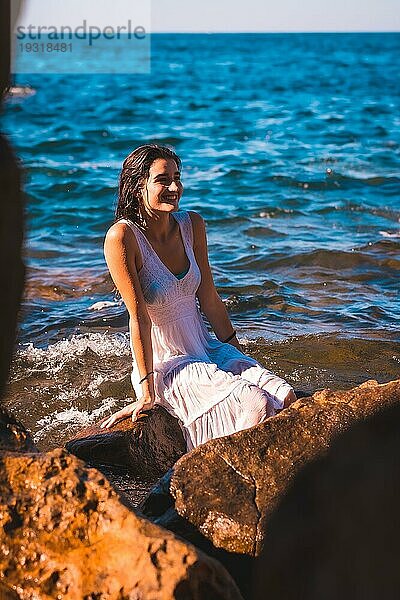 Ein junges kaukasisches Mädchen in einem nassen weißen Kleid sitzt lächelnd auf einem Felsen am Meer