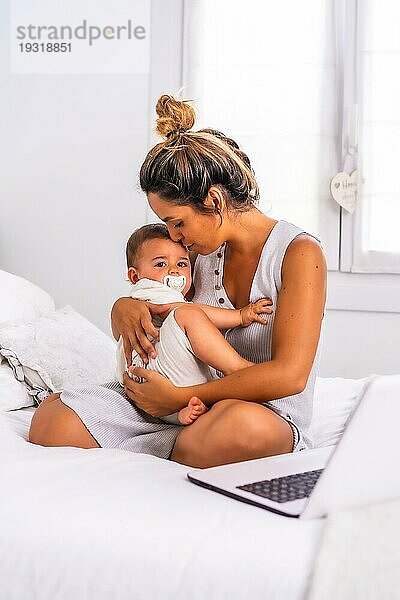 Junge kaukasische Mutter mit ihrem Sohn im Zimmer oben auf dem Bett. Baby weniger als ein Jahr lernen die ersten Lektionen ihrer Mutter. Umarmen ihn und geben ihm Liebe