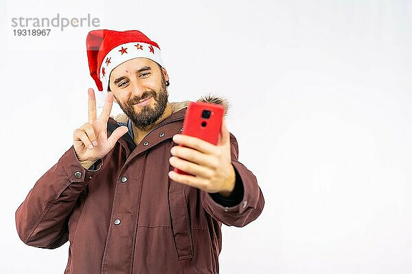 Sehr glücklicher junger kaukasischer Mann mit roter Weihnachtsmütze  der ein Selfie auf weißem Hintergrund macht  bekleidet mit einer Winterjacke