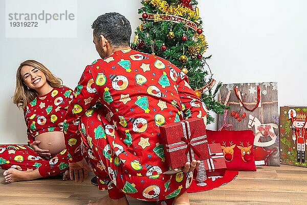 Junges Paar sitzt am Weihnachtsbaum  der Freund schenkt seiner Freundin lächelnd ein Geschenk