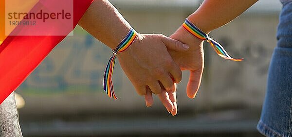 Eine Nahaufnahme von zwei jungen kaukasischen Frauen  die sich mit LGBT Stolz Armbändern im Freien an den Händen halten