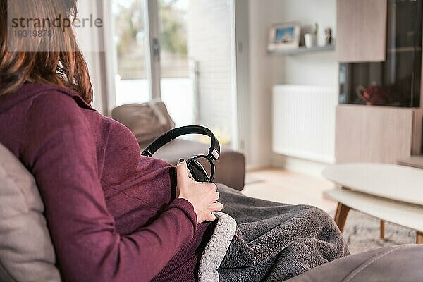 Detail einer jungen schwangeren kaukasischen Frau  die sich mit ihrem Kind verbindet  indem sie mit Kopfhörern Musik auflegt  in der Wohlfühlatmosphäre ihres Hauses