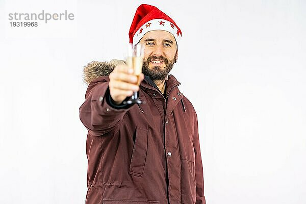 Junger  sehr glücklicher kaukasischer Mann mit roter Weihnachtsmütze  der mit einem Glas Champagner auf einem weißen Hintergrund anstößt  gekleidet in eine Winterjacke