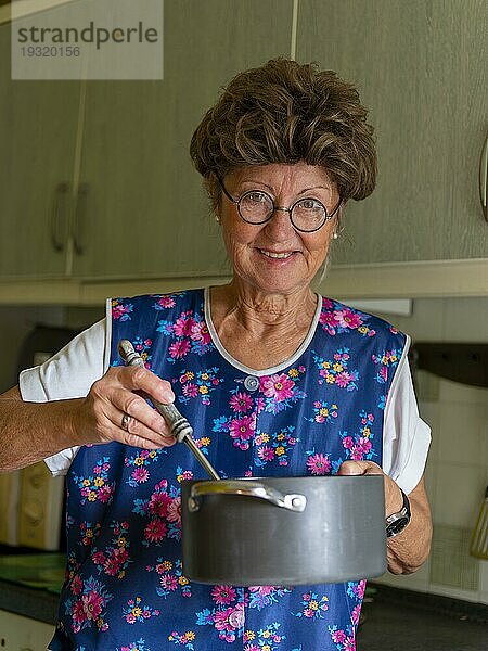 Oma mit alter Kittelschürze  Brille und Perücke in der Küche  hält Kochtopf und Kelle in Händen  Deutschland  Europa