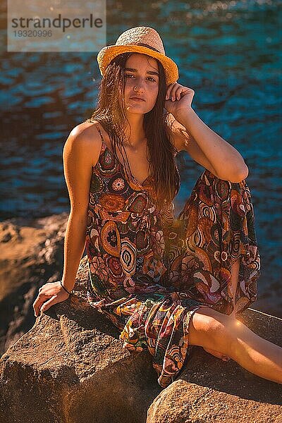 Blick einer jungen kaukasischen Frau mit nassen Haaren  einem geblümten Kleid und einem Hut  die auf einem Felsen am Meer sitzt  Sommerleben in der goldenen Stunde