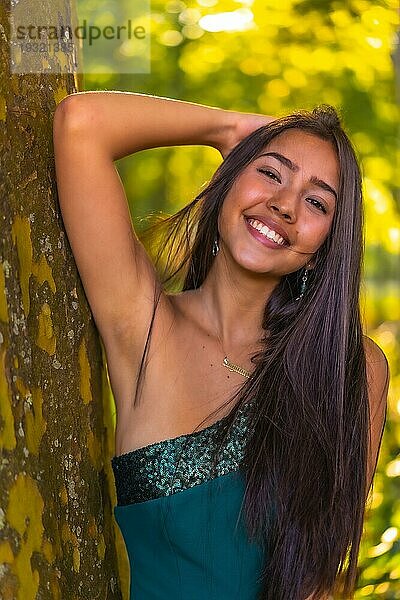 Eine junge brünette Latina mit langen glatten Haaren lehnt in einem grünen Kleid an einem Baum. Porträt einer nachdenklichen und sinnlichen jungen Frau  vertikales Foto