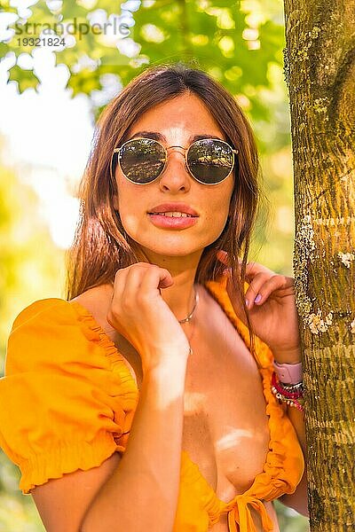 Lifestyle einer jungen kaukasischen jungen Brünette in einem Park im Sommer neben einem Baum in der Natur. Mädchen mit Brille lächelnd  Blick in die Kamera