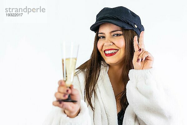 Junges lateinamerikanisches Mädchen mit schwarzem Hut feiert Silvester auf einem weißen Hintergrund. Porträt mit einem Glas Champagner