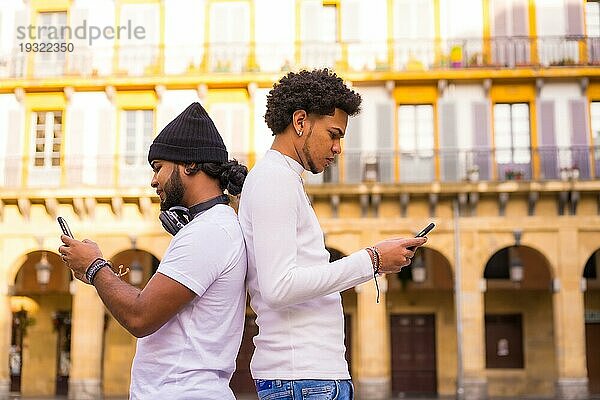 Lifestyle  zwei schwarze Latino Freunde  die Nachrichten versenden oder soziale Netzwerke auf ihrem Handy beobachten. Junge mit Afrohaar und Junge mit Hut und Kopfhörern