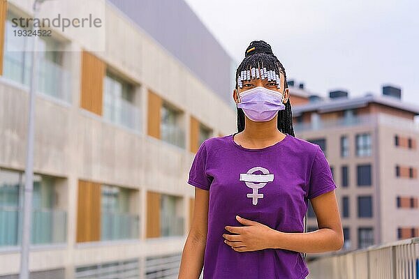 Internationaler Frauentag am 8. März im Jahr der Coronaviruspandemie Covid 19. Dominikanische Frau mit Zöpfen und feministischem lila TShirt und Gesichtsmaske