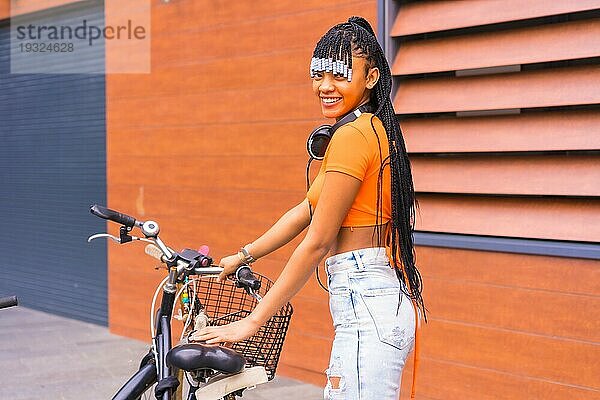 Lifestyle mit einer jungen Trap Tänzerin in der Stadt. Schwarzes Mädchen der afrikanischen ethnischen Gruppe mit orangefarbenem Hemd in der Stadt. Cofing Fahrrad geparkt in der Stadt  lächelnd