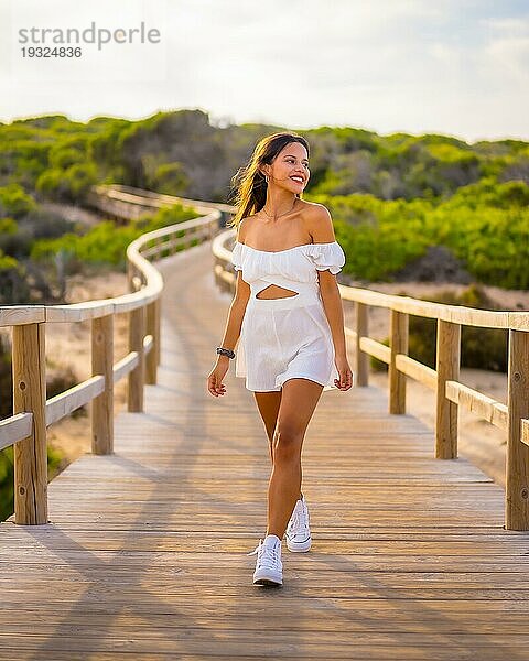 Lebensstil einer jungen brünetten Kaukasierin  die im Sommer in einem weißen Kleid den Strandurlaub genießt und den Holzsteg entlang geht
