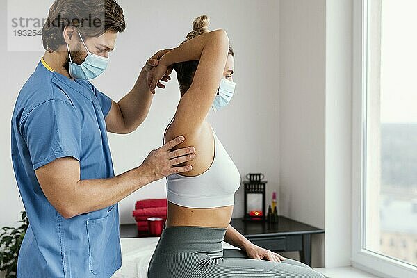 Osteopathischer Therapeut mit medizinischer Maske prüft das Schultergelenk einer Patientin