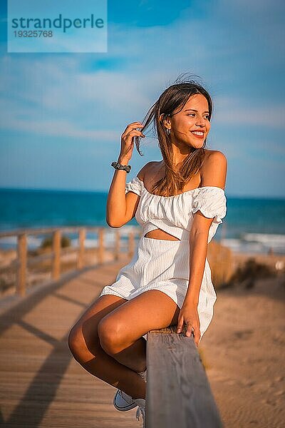 Lebensstil einer jungen brünetten Kaukasierin im Urlaub am Strand in einem weißen Kleid im Sommer  sitzend auf dem Holzsteg