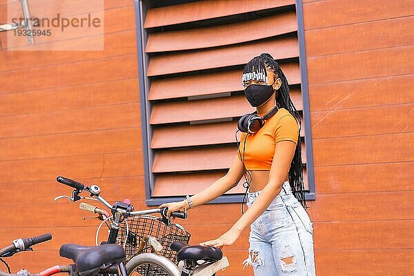 Schwarze Rasse Mädchen mit Maske von Coronavirus Pandemie  afrikanische ethnische Gruppe mit orangefarbenen Hemd in der Stadt. Cofing das Fahrrad in der Stadt geparkt