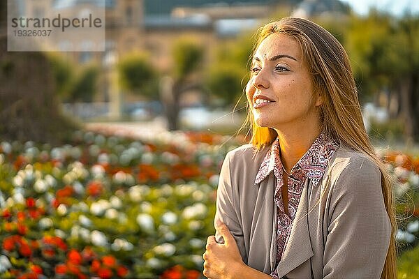 Lifestyle  ein junger Unternehmer. Kaukasierin und Blondine mit kastanienbraunem Regenmantel und Blumenkleid in der Stadt. Lächelnde Schicht neben Blumen in einem Park mit Blick nach links